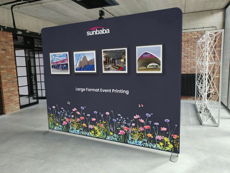 Sunbaba branded zipper wall display in a modern industrial-style room, featuring framed images of event setups and floral graphics along the bottom with the text 'Large Format Event Printing'